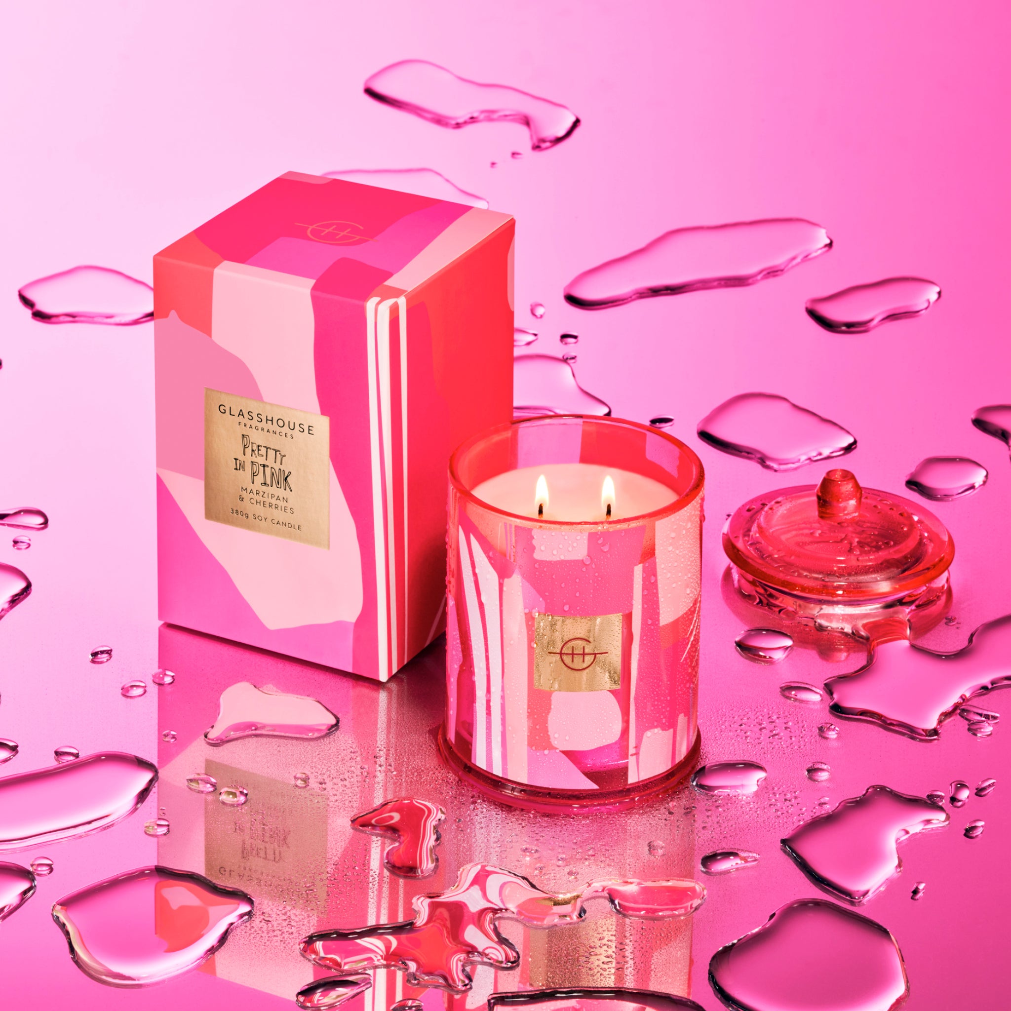 Glasshouse Fragrances Pretty in Pink Marzipan and Rhubarb 380g Soy Candle burning beside box on a wet pink mirror surface