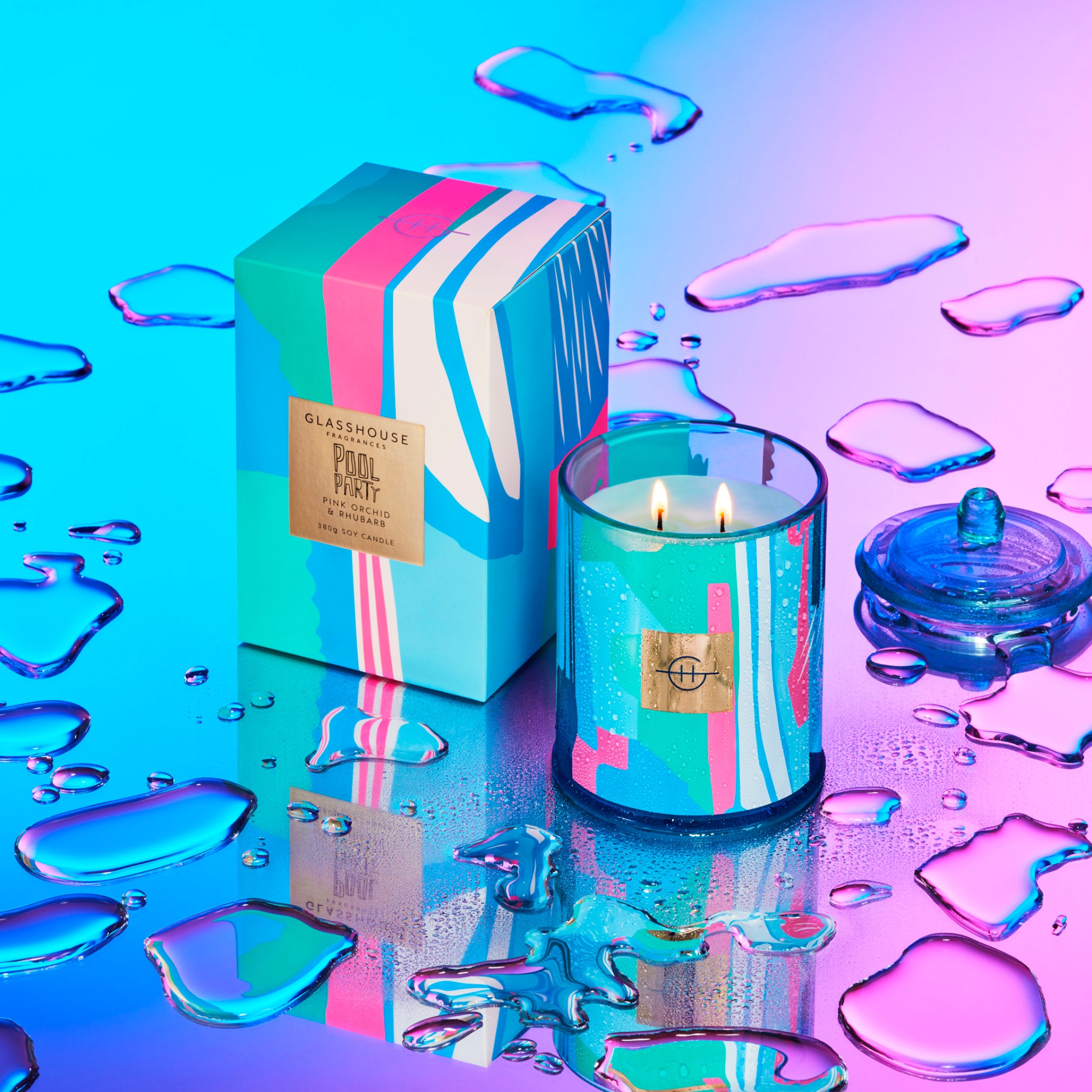 Glasshouse Fragrances Pool Party Pink Orchid and Rhubarb 380g Soy Candle burning beside box on a wet colourful mirror surface